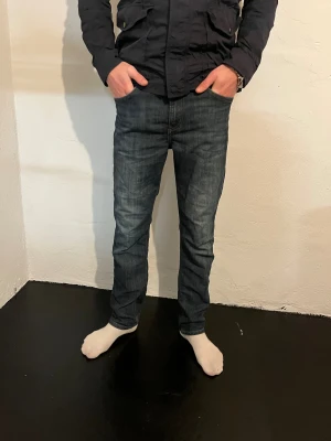 Mörkblå jeans från Lee - Säljer ett par mörkblå jeans från Lee med klassisk femficksdesign och snygga slitningar. Jeansen har raka ben och normal passform, med Lee-logga på bakfickan och läderpatch i midjan. Perfekta för dig som gillar en tidlös och avslappnad stil. 