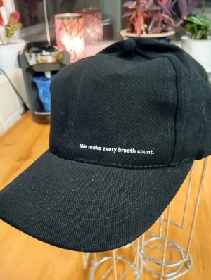 Svart keps med text framtill - Stilren svart keps med böjd skärm och vit text 'We make every breath count.' framtill. Tillverkad i ett slitstarkt bomullsmaterial som ger en klassisk look. Perfekt för dig som gillar enkel och clean streetstyle.