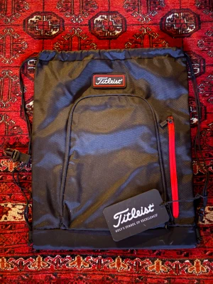 Svart ryggsäck från Titleist med röd dragkedja - Snygg svart ryggsäck från Titleist med röd dragkedja på framsidan och praktisk snörning upptill. Väskan är tillverkad i slitstark polyester och har ett stilrent Titleist-märke. Perfekt för att bära med sig grejer till träningen eller skolan.