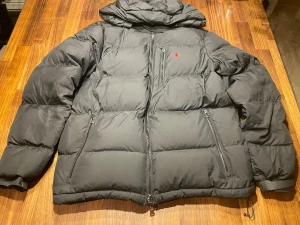 Svart dunjacka från Polo Ralph Lauren - Svart dunjacka från Polo Ralph Lauren med huva, flera dragkedjefickor och röd broderad logga på bröstet. Jackan är quiltad och har en loose passform, perfekt för kalla dagar. Materialet är polyester och dun, vilket gör den både varm och lätt.