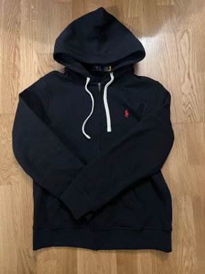 Svart hoodie från Polo Ralph Lauren - Svart hoodie från Polo Ralph Lauren med vit snörning och dragkedja framtill. Klassisk röd Polo-logga på bröstet och stor huva. Tröjan har långa ärmar och två fickor framtill. 
