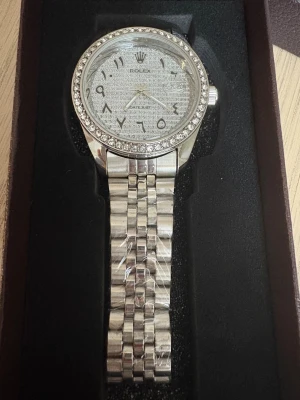 Roli Datejust silver  - Snygg Roli Datejust med silverfärgat armband i metall och glittrande stenar runt urtavlan. Urtavlan har ett mönstrat motiv och arabiska siffror, samt en klassisk Roli-logga. 