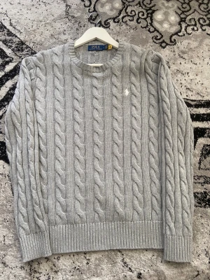 Grå kabelstickad tröja Polo Ralph Lauren - Snygg grå kabelstickad tröja från Polo Ralph Lauren i storlek M. Tröjan har rund hals, ribbade muddar och ett diskret broderat logomärke på bröstet. Perfekt för dig som gillar klassisk och stilren design med en modern touch.