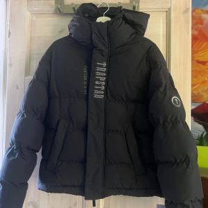 Svart pufferjacka från Trapstar - Svart pufferjacka från Trapstar med stor broderad logga och texten 'IT'S A SECRET' på både fram- och baksidan. Jackan har huva, två sidofickor och en chunky look. Materialet är syntet och jackan är perfekt för kalla dagar. Mycket fint skick, använd fåtal gånger så som ny utan prislapp. Finns inte att köpa längre och jag betalde 5000 kr, plus frakt då den är köpt via Trapstar uk. Pris kan diskuteras! 
