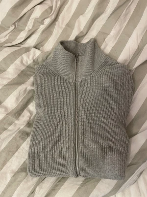 Grå stickad kofta från East West - Snygg grå stickad kofta från East West med ribbad struktur och hel dragkedja framtill. Hög krage och långärmad modell som ger en clean och chill vibe. Perfekt att slänga på sig när det är lite kyligt ute.