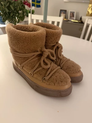 Beiga inuikiis i teddy och skinn - Säljer ett par beiga inuikiis med fluffig teddytextur och detaljer i skinn. Skorna har snörning runt hela skaftet och en platt sula. Perfekta för kalla dagar när du vill ha både stil och värme. Endast använda fåtal gånger.