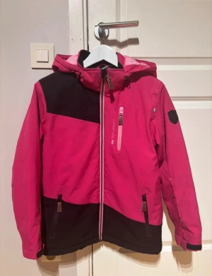 Rosa och svart vinterjacka  - Snygg vindjacka från Lindberg i rosa och svart med huva och flera dragkedjor. Jackan har lång ärm, justerbara muddar och coolt snitt med diagonal färgblock. Perfekt för dig under vinterns kyliga dagar 🩵🌨️☃️