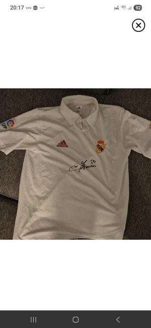 Signerad Real Madrid tröja av lilian thuram. - Säljer en signerad vit orginal Real Madrid jubileumströja från Adidas, storlek M. Tröjan har korta ärmar, klassisk krage och broderade detaljer med klubbmärke och jubileum 1902-2002. LFP-logga på ärmen och autograf på bröstet av Lilian Thuram.