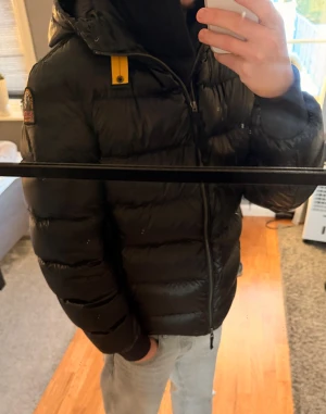 Svart dunjacka från Parajumpers - Säljer en svart dunjacka från Parajumpers med huva och gul detalj vid kragen. Jackan har ett snyggt, puffigt quiltat utseende, ribbade muddar och dragkedja. Har storleken L men sitter som M. Använd en vinter och i topp skick.
