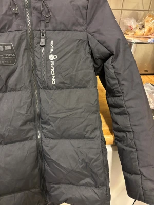 Svart Sail Racing dunjacka S - Svart dunjacka från Sail Racing med huva och coola loggor på bröstet, ärmen och ryggen. Jackan har dragkedja framtill, flera fickor och quiltad design. Perfekt för kalla dagar och har en schysst, sportig look.