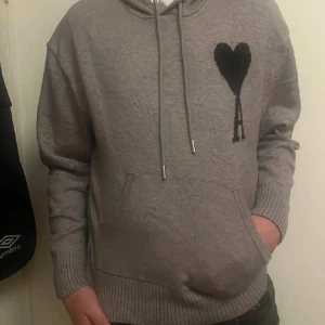Ami Paris hoodie - Snygg grå hoodie från Ami Alexandre Mattiussi med svart hjärta och bokstaven A broderat på bröstet. Tröjan har huva med snörning, ribbade muddar och magficka. Perfekt för dig som gillar stilrena detaljer och vill ha en chill look.
