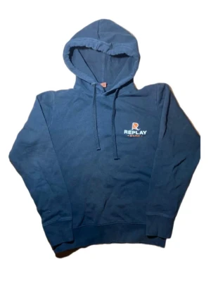 Blå hoodie från Replay med huva - Säljer en snygg blå hoodie från Replay med tryckt logga på bröstet. Tröjan har huva med snörning och långa ärmar, perfekt för chill dagar. Mjuk bomullskvalitet och ribbade muddar vid ärmslut och nederkant. Passar dig som gillar streetwear.