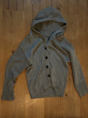 Grå kofta med huva och knappar - Snygg grå kofta med huva och svarta knappar framtill. Tröjan är stickad i mjukt material och har långa ärmar med ribbade muddar. Perfekt för dig som gillar chill och enkel stil. Passar dig som vill ha en normal passform och gillar klassiska detaljer.