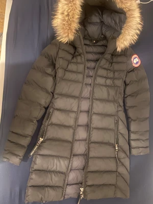 Svart lång dunjacka Canada Goose - Säljer en svart lång dunjacka från Canada Goose med fluffig beige päls på huvan. Jackan har två dragkedjefickor, quiltad design och klassisk patch på ärmen. Perfekt för kalla vinterdagar och har en snygg, stilren look.