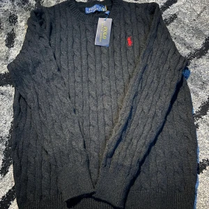 Svart kabelstickad tröja Polo Ralph Lauren - Svart kabelstickad tröja från Polo Ralph Lauren med klassisk rund hals och röd broderad logga på bröstet. Tröjan har långa ärmar och ribbade muddar vid ärmslut och nederkant. Perfekt för dig som gillar stilren och tidlös design.