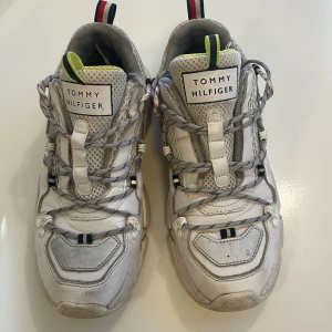 Chunky sneakers från Tommy Hilfiger - Chunky vita sneakers från Tommy Hilfiger med grå och röda detaljer. Skorna har meshpaneler, snörning och en robust sula. Detaljer i neon och klassiska Hilfiger-färger på hälflikarna. Perfekta för en sportig och trendig look.