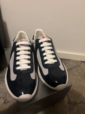 Prada cups  - Snygga Prada sneakers med marinblå lackdetaljer och vit mesh. Klassisk rund tå, vita snören och vit platt sula. Röd Prada-logga på plösen ger en exklusiv touch. Perfekt för dig som gillar stilrena och sportiga designer.