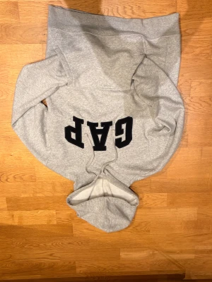 Grå hoodie från GAP med logga - Säljer en grå hoodie från GAP i storlek XS/s. Tröjan har en stor svart GAP-logga på bröstet, huva med snörning och långa ärmar. Mjuk insida och klassisk passform, perfekt för chill dagar. Materialet är bomull och polyester.
