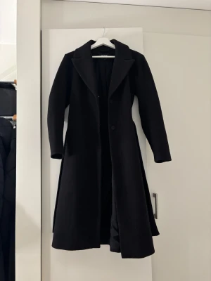 Svart lång kappa från ONLY XS - Säljer en svart lång kappa från ONLY i storlek XS. Den har klassisk krage, knappar framtill och ett matchande bälte i midjan. Kappan är stilren och har en snygg, rak passform med långa ärmar. Perfekt för dig som gillar minimalistisk och clean look.