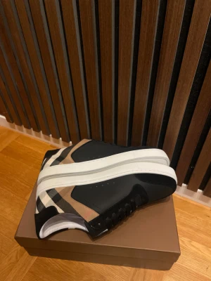 Burberry rutig sneakers med svart skinn - Snygga Burberry sneakers med klassiskt rutig mönster i beige, svart och vitt. Skorna har svarta skinnpaneler, vita platta sulor och svarta snören. Perfekt för dig som gillar lyxiga detaljer och ikonisk design. Kommer med originalkartong och dustbag.
