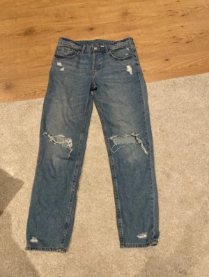 Blå slitna jeans från H&M Divided - Säljer ett par blå jeans från H&M Divided i storlek 36. Jeansen har raka ben, slitningar och hål på knäna samt en broderad figur på ena bakfickan. Klassisk femficksmodell i denim med normal passform.