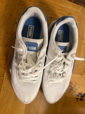 Vita och blå sneakers från Puma - Säljer ett par fräscha vita sneakers från Puma med blå detaljer och klassisk logga på sidan och plösen. Skorna har snörning, platt sula med bra grepp och är tillverkade i syntetmaterial. Perfekta för dig som gillar sportig och clean stil.
