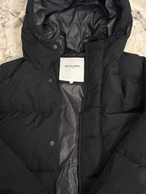 Svart pufferjacka från Jack & Jones - Svart pufferjacka från Jack & Jones med huva och dragkedja framtill. Jackan har en clean look med slät yta och fodrad insida i mörkgrått. Perfekt för kalla dagar och enkel att matcha med olika outfits.