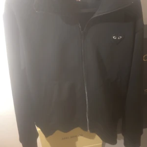 Svart zipjacka från Comme des Garçons - Svart zipjacka från Comme des Garçons Play med ikoniskt hjärta med ögon på bröstet och stort tryck på ryggen. Jackan har hög krage, dragkedja framtill och två fickor. Perfekt för dig som gillar streetwear och vill sticka ut med en cool detalj.