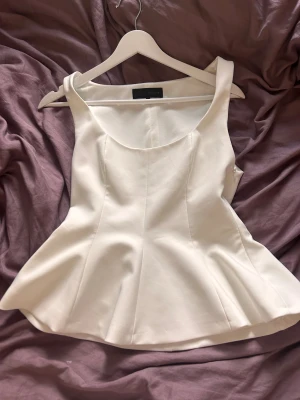 Vit peplumtop från Zara - Supersnygg vit peplumtop från Zara med breda axelband och rundad halsringning. Toppen har en figurnära passform upptill och ett voluminöst peplum nedtill som ger en cool siluett. Materialet är slätt och har lite glans, perfekt att styla med jeans eller kjol.