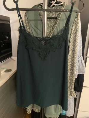 Mörkgrön linnetopp med spets  - Supersnygg mörkgrön linnetopp med tunna axelband och v-ringad spetskant framtill. Perfekt att styla med jeans eller kjol för en cool look. Materialet är mjukt och lätt, och spetsen ger en extra touch av elegans.