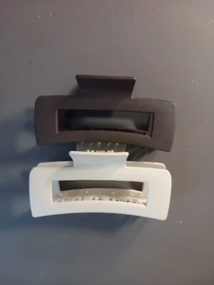 Två stora hårklämmor i plast - Säljer två rektangulära hårklämmor i plast, en i matt brun och en i matt vit. Båda har stilren, minimalistisk design med bred öppning och stark fjäder. Perfekta för att snabbt sätta upp håret i en snygg look. Från Gina Tricot, andvänd en del men fortfarande fint skick!