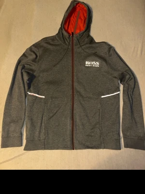 Grå zip hoodie från Hugo Boss - Snygg grå hoodie från Hugo Boss med dragkedja och vit logga på bröstet. Hoodien har huva med orange insida och dragsnören, samt två fickor med vita detaljer. Perfekt för en avslappnad och stilren look.