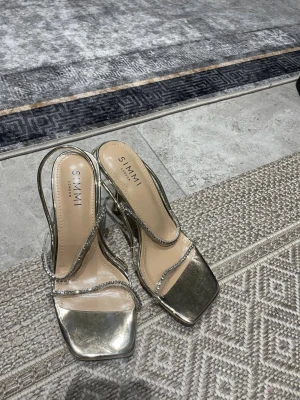 Guldiga pumps med strass från Simmi - Säljer ett par glammiga guldiga pumps från Simmi London med fyrkantig tå och smala remmar dekorerade med strass. Skorna har hög klack och öppen häl, perfekta för att lyfta vilken outfit som helst.