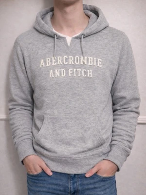 Grå hoodie från Abercrombie & Fitch - Snygg grå hoodie från Abercrombie & Fitch med broderad logga på bröstet. Klassisk känguruficka framtill, dragsko i huvan och mjukt material som känns skönt mot huden. Perfekt för en avslappnad och trendig look.