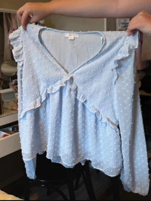 Ljusblå volangblus med prickar - Supersöt ljusblå blus med volangdetaljer och små prickar i strukturen. Djup V-ringning framtill och långa ärmar med volang vid axlarna. Blusen har en lös passform och lätt rynk vid midjan för en feminin siluett. Jättebra skick o från Gina i storlek 170