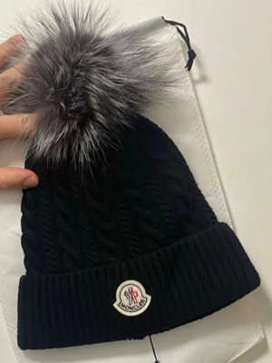 Svart Moncler mössa med pälsboll - Snygg svart stickad Moncler mössa med stor fluffig pälsboll i grått och svart. Klassisk Moncler-logga framtill och bred vikt kant. Perfekt accessoar för kalla dagar och ger en lyxig touch till din outfit.