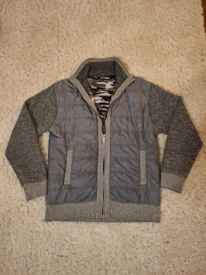 Grå stickad tröja med quiltad front - Snygg grå tröja med stickade ärmar och rygg samt quiltad front. Tröjan har dragkedja, hög krage och två fickor framtill. Perfekt för lager-på-lager och ger en cool, urban look. Melerad grå färg och mjukt material.