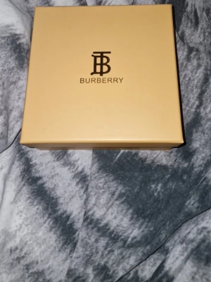 Burberry bälte med klassiskt rutmönster - Snyggt Burberry bälte med det ikoniska beige rutmönstret och svarta kanter. Bältet har ett stilrent metallspänne och levereras i originalask. Perfekt accessoar för att lyfta din outfit med en lyxig touch.