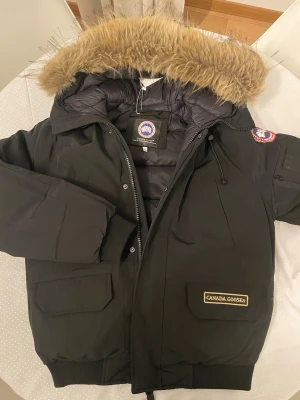 Svart parkas från Canada Goose - Säljer en svart parkas från Canada Goose med stor huva och fluffig pälsimitation. Jackan har flera fickor, patch på ärmen och dragkedja med vindslå. Perfekt för kalla dagar och riktigt snygg streetstil.