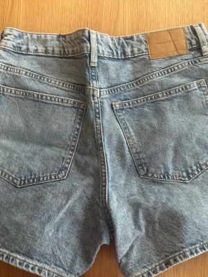Blå denimshorts från hm - Klassiska blå jeansshorts från hm i mjuk bomullsdenim. Modellen har fem fickor, bälteshällor och knappgylf. Perfekta för sommaren och har en avslappnad passform med något högre midja.