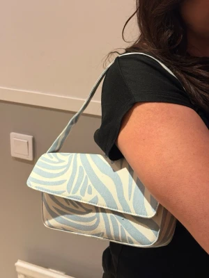 Blåvit zebramönstrad handväska - Säljer en trendig handväska i blått och vitt med zebramönster. Väskan har en rektangulär form och axelrem, perfekt för att bära det viktigaste. Haft i flera år men aldrig använd, så väskan är precis som ny.  Priset går att förhandla! 
