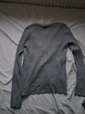 Grå ribbstickad tröja med spetskant - Grå långärmad ribbstickad tröja med fin spetsdetalj runt halsringningen. Tröjan har en smal passform och är perfekt för lager-på-lager. Enkel och stilren design som passar till det mesta.