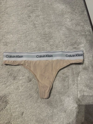 Beige stringtrosa från Calvin Klein - Snygg beige stringtrosa från Calvin Klein med bred resår i midjan där loggan syns tydligt. Trosan har en klassisk stringmodell och är tillverkad i mjukt och stretchigt material för skön passform. Perfekt för dig som gillar stilrena och bekväma underkläder.