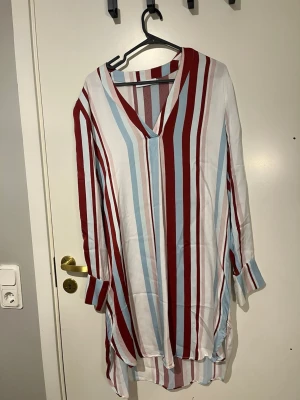 Randig klänning med v-ringning - Säljer en lös och luftig klänning med v-ringning och långa ärmar. Klänningen har vertikala ränder i rött, vitt och ljusblått. Använd en gång.