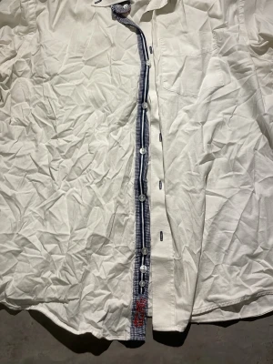 Vit skjorta från Storm Copenhagen - Säljer en vit skjorta från Storm Copenhagen med blåvitrutiga detaljer längs knappslån och insidan av kragen. Skjortan har klassisk krage, bröstficka och knappar framtill. Perfekt för dig som gillar stilrena plagg med en twist.