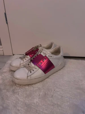 Valentino Open sneakers med rosa detalj - Snygga vita Valentino Open sneakers med en bred metallicrosa rem över vristen. Skorna har klassisk rund tå, snörning och är tillverkade i skinn. Perfekta för dig som vill sticka ut med en färgklick i din outfit. Skriv vid fler frågor osv
