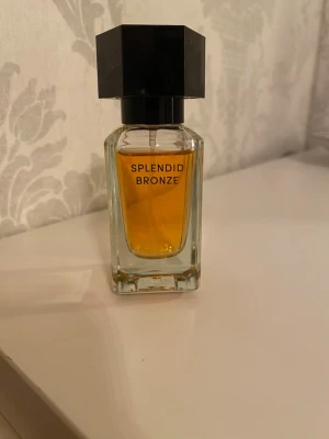 Splendid Bronze parfym - En elegant parfym från Splendid Bronze i en rektangulär glasflaska med svart, sexkantigt lock. Flaskan har en klar design och innehållet är bärnstensfärgat vilket ger ett lyxigt intryck. Perfekt för dig som gillar stilrena och moderna dofter.