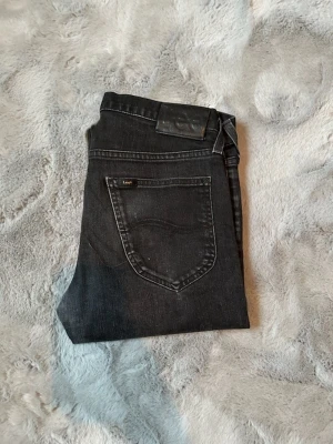 Svarta jeans från Lee - Säljer ett par svarta jeans I storlek W30 L32 från Lee med klassisk femficksdesign och Lee-logga på bakfickan. Jeansen har en bra passform och är tillverkade i mjukt denimtyg som sitter snyggt. Perfekta för dig som gillar en stilren och enkel look.