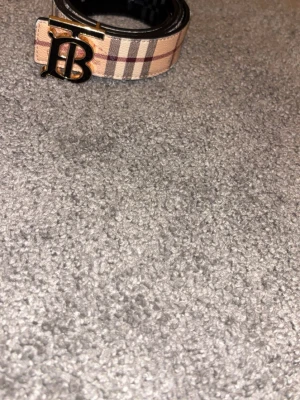 Burberry bälte med rutigt mönster - Snyggt Burberry bälte med klassiskt beige och brunt rutmönster på ena sidan och svart läder på andra sidan. Spännet är i metall med Burberrys ikoniska TB-logga. Perfekt accessoar för att lyfta vilken outfit som helst.