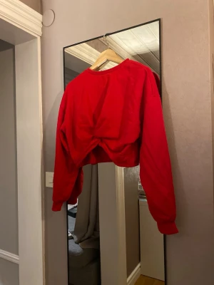 Röd croppad sweatshirt med twist - Trendig röd croppad sweatshirt med twistad detalj framtill. Tröjan har rund halsringning och långa ärmar med avslappnad passform. Perfekt för en cool och avslappnad stil. Materialet ser ut att vara mjuk bomullsblandning.